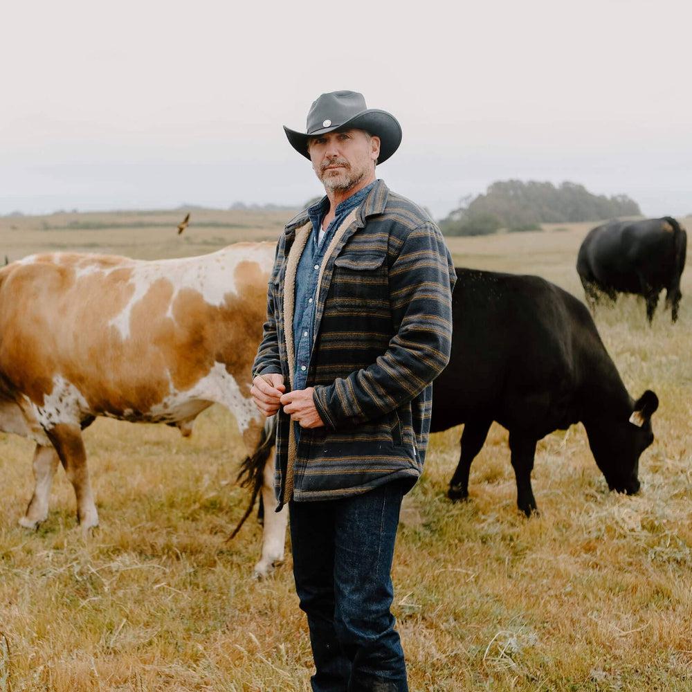 A man with cows behind him wearing Cyclone Black Leather Cowboy Hat with 3" Brim and 4" Crown