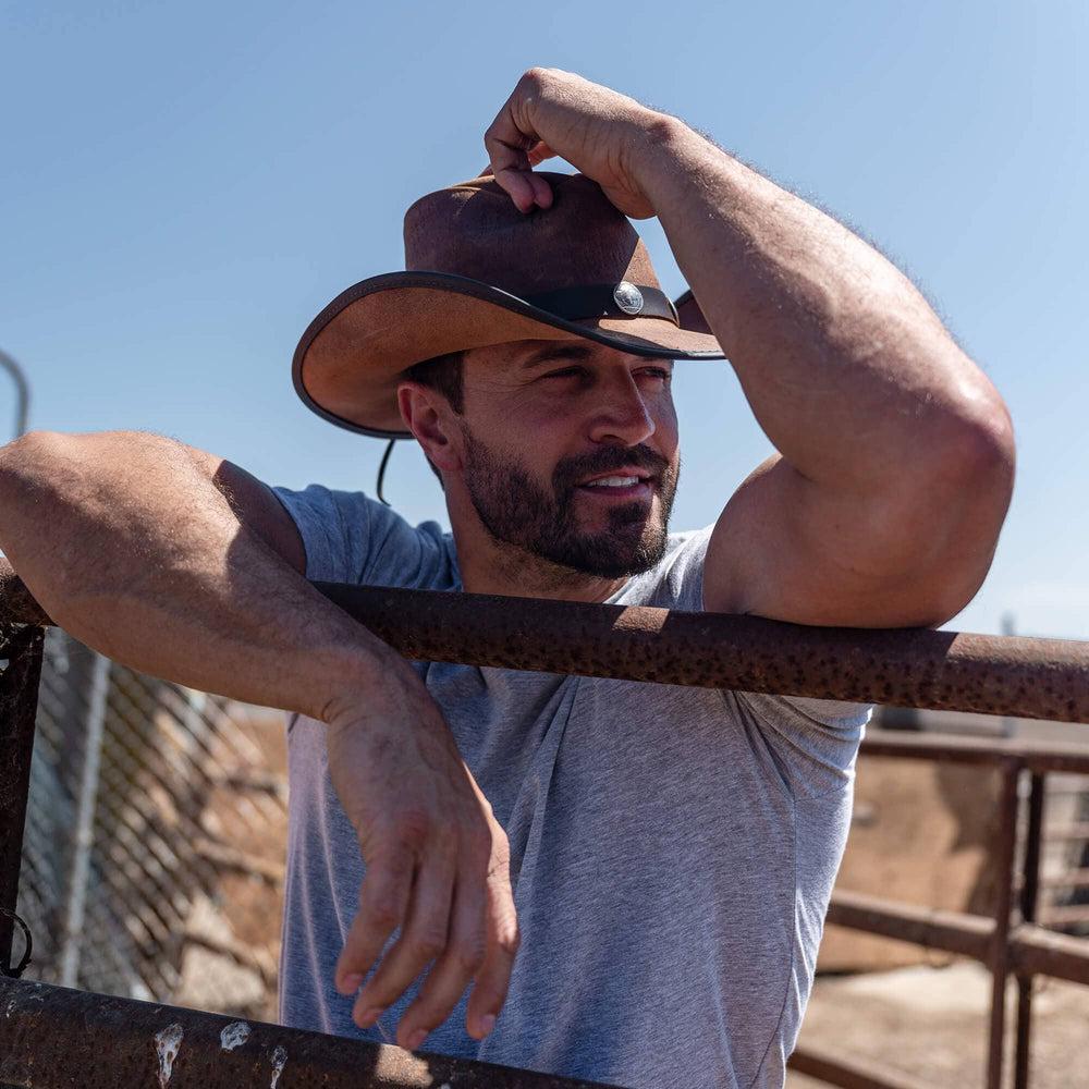 A man leaning on a steel fence wearing leader cowboy hat