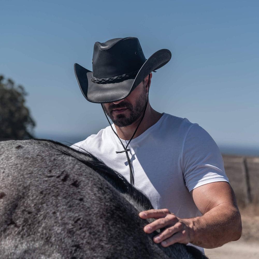 A man wearing a leather cowboy hat touching a horse