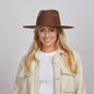 A woman with long blonde hair, smiling and wearing a brown Rancher hat and a textured cream coat over a white shirt