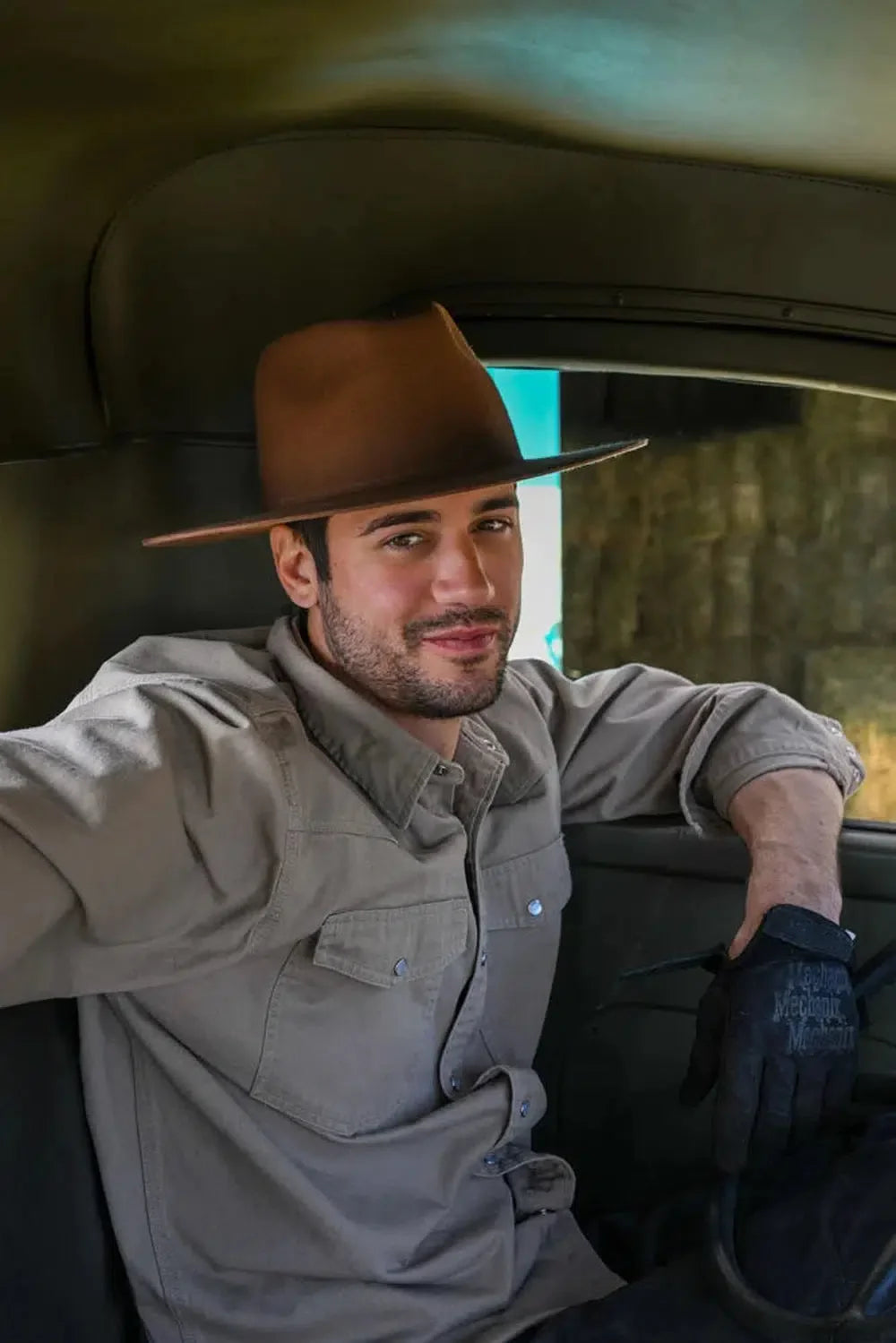 A man sitting inside a truck wearing a gray polo, gloves and brown felt hat