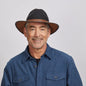 A man with a light stubble wearing a felt coal fedora hat and a gray button-up shirt.