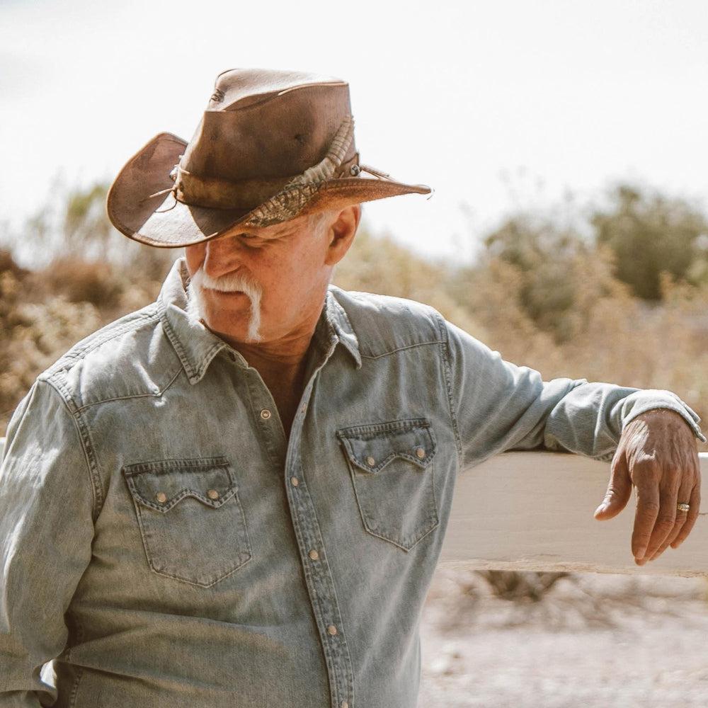 A man leaning on a wooden fence wearing a denim jacket and a cowboy hat